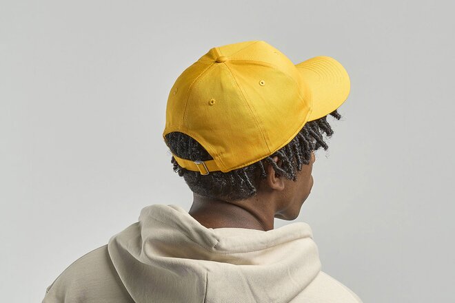 Mann iført en gul caps med mesh-paneler og justerbar stropp. Han har på seg en beige hettegenser. Capsens pustende netting gir en luftig og komfortabel passform.