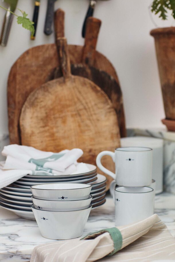 Hvitt Lexington-servise med stjernedetaljer, stablet sammen med kjøkkenhåndklær og tre skjærebrett i et rustikt kjøkkenmiljø.