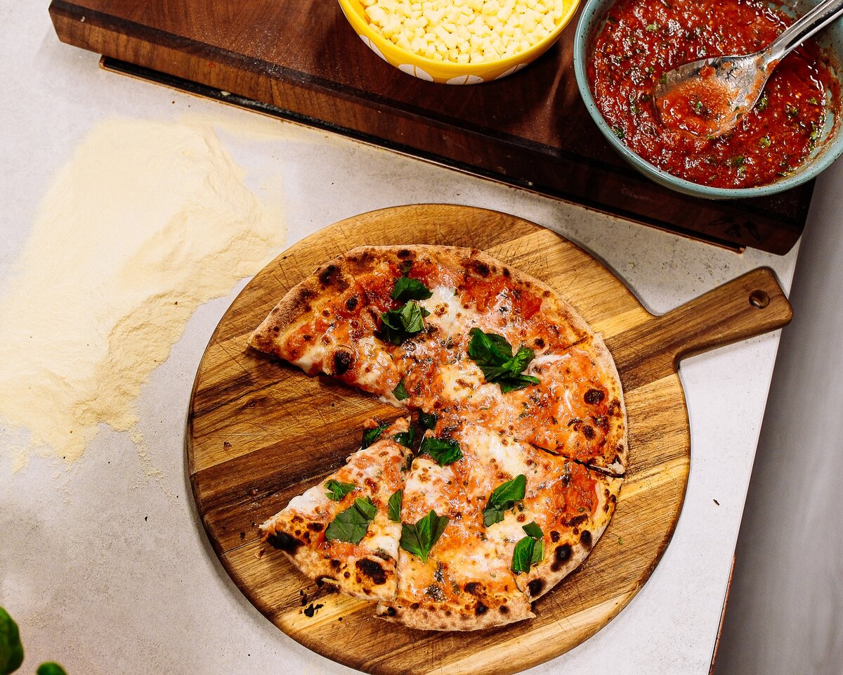 Nybakt pizza margherita med basilikum på en rund treplate, med revet ost og tomatsaus i boller ved siden av.