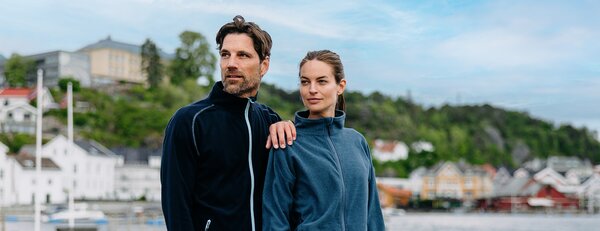 Mann og kvinne iført fleecejakker fra YOU Brands i maritimt miljø med norsk kystlandskap i bakgrunnen.