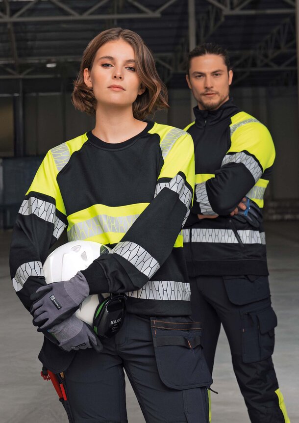 En kvinne og en mann iført gul-svart Jobman-refleksklær, stående i en industribygning med verneutstyr.