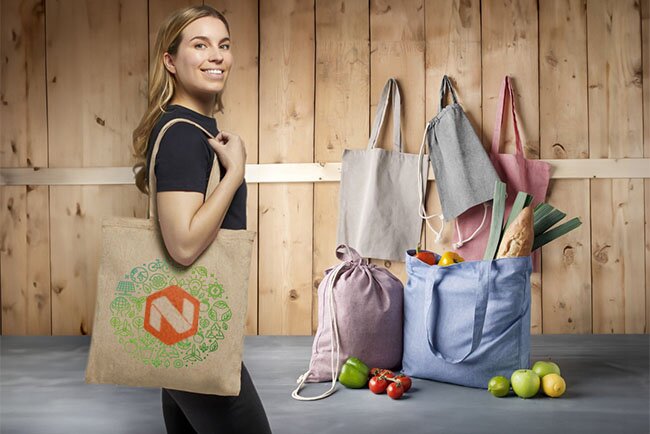 Kvinne med et miljøvennlig handlenett laget av jute, med trykt logo og bærekraftig design. Flere bærenett i bakgrunnen fylt med dagligvarer.