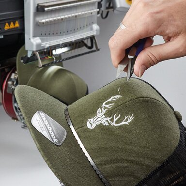 Detaljarbeid på brodert logo på olivengrønn caps. En hånd bruker et verktøy for å fjerne overskuddstråder fra broderiet.