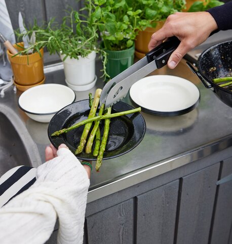 Sagaform grilltang i bruk for å servere grillede asparges på en svart emaljert Sagaform tallerken, med friske urter og servise i bakgrunnen. Perfekt for grilling og utendørs matlaging.