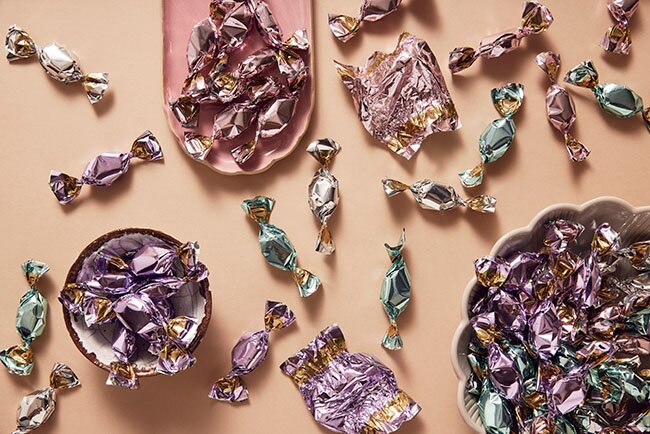 Små sjokoladebiter i glitrende innpakning i rosa, lilla og blå toner, spredt på en beige bakgrunn. Perfekte giveaways for messer, events og promotering.