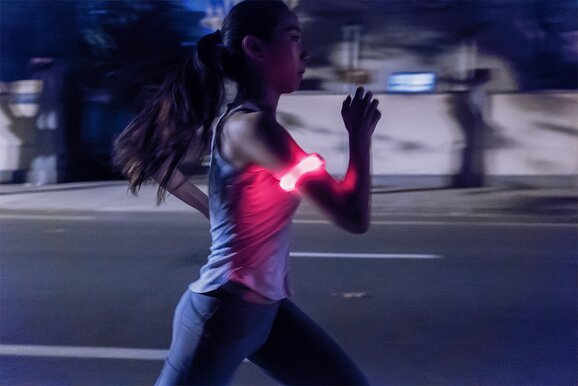 Kvinne jogger i mørket iført et refleksbånd med røde lysende led lys på overarmen. Refleksen gir økt synlighet i trafikken og bidrar til bedre sikkerhet for mosjonister. Perfekt som profilartikkel for reflekskampanjer.