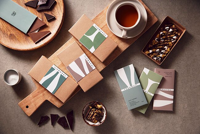 Høykvalitets sjokolade med profilert innpakning, presentert på et trefat med kaffe og nøtter. Perfekt profileringssjokolade for firmagaver og kampanjer.