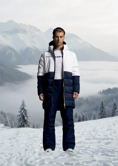 Mann iført hvit og mørkeblå Craft-dunjakke, stående i et snødekt fjellandskap.
