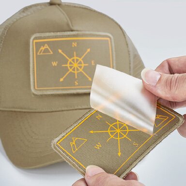 Stoffmerke med trykt broderi-effekt på caps. En hånd fjerner overføringsfolien fra merket før det monteres på capsen. Motivet er varmeoverført og designet for å ligne broderi.