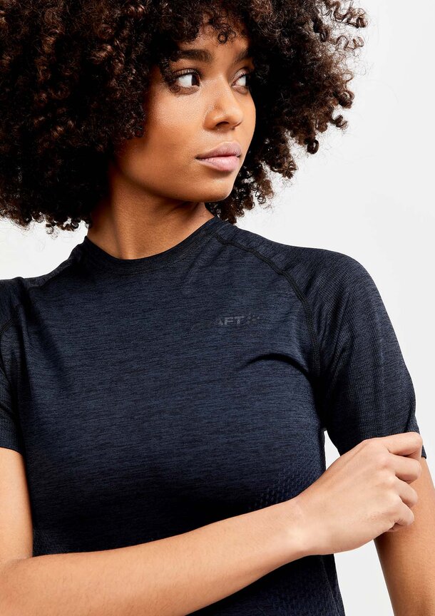 Kvinne med krøllet hår iført en mørk Craft Teamwear-T-skjorte, sett fra overkroppen og opp.