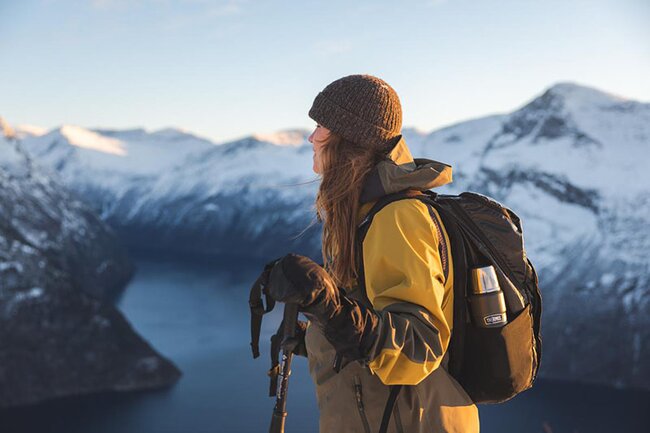 Tur og fritid – kvinne med tursekk og termos nyter utsikt over snødekte fjell og fjord