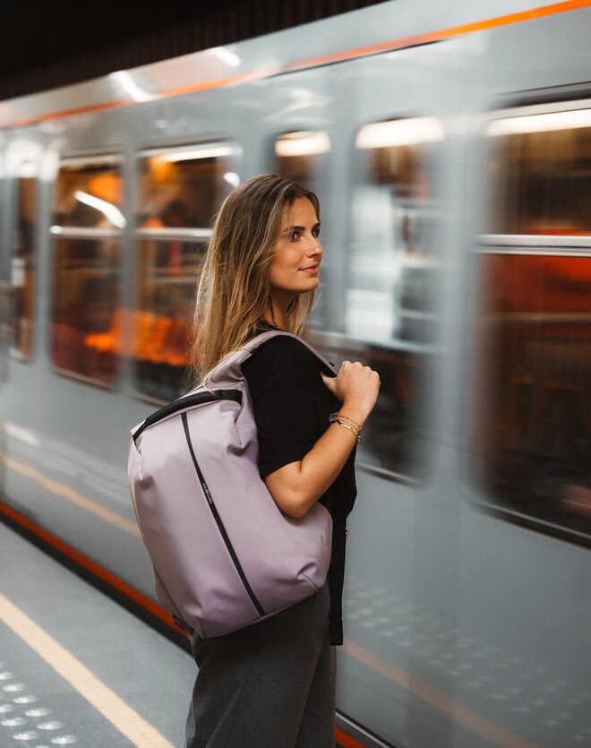 Moderne ryggsekk i minimalistisk design båret av en kvinne på en travel togstasjon. Den stilrene sekken kombinerer funksjonalitet og urban stil, perfekt for pendling, jobb og reiser. Lettvektsmateriale og ergonomiske skulderstropper gir komfort hele dagen.