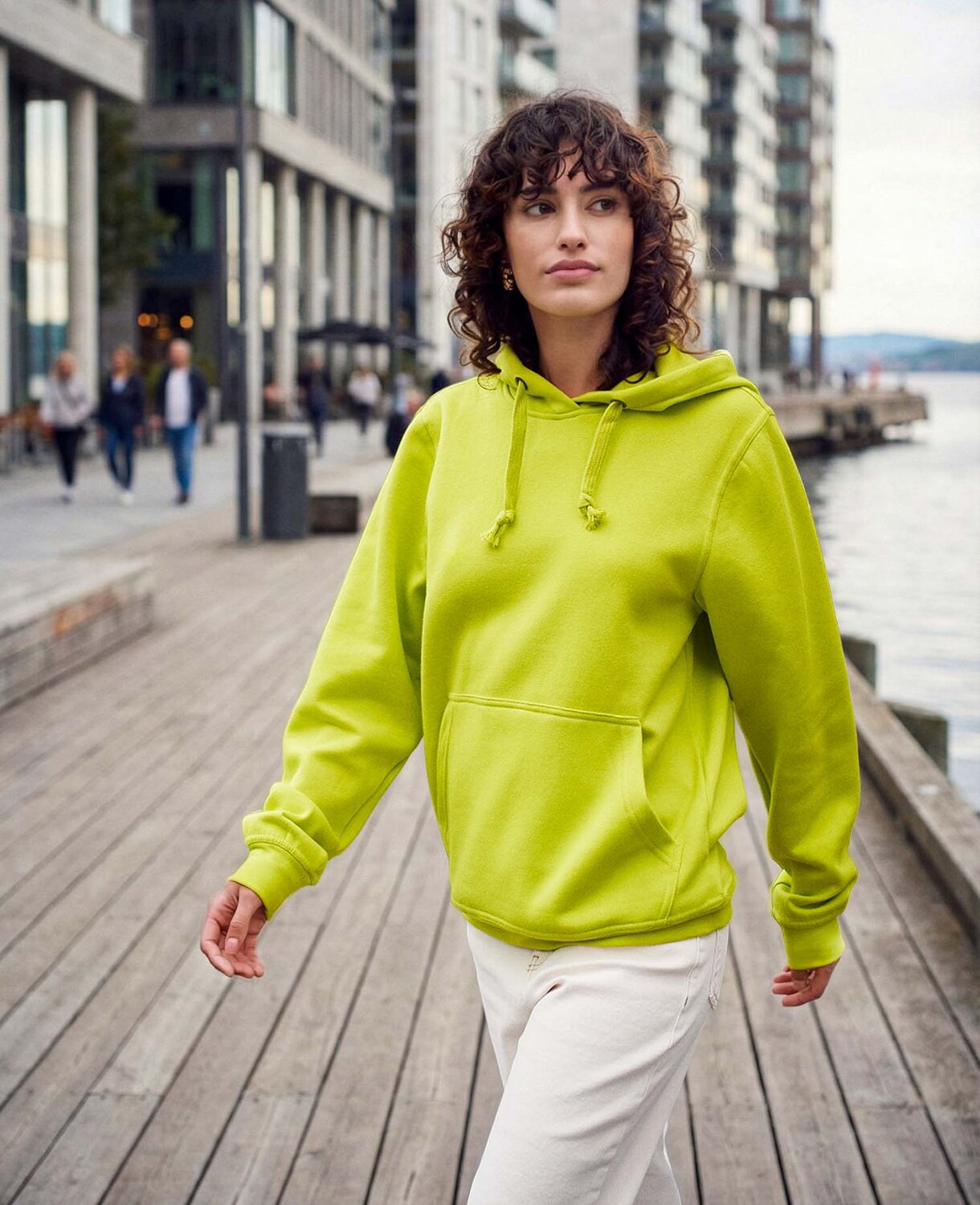 Modell i limegrønn hettegenser som går langs brygge i urbant miljø – unisex hoodie i bomull med kengurulomme og snøring.
