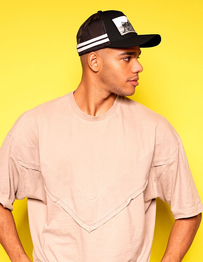 Mann iført en svart og hvit trucker-caps med trykt logo på frontpanelet. Capsen har mesh-paneler bak og buet brem. Han har på seg en beige oversized t-skjorte og står mot en gul bakgrunn, vendt til siden.