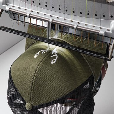 Grønn og sort mesh-caps i ferd med å bli brodert med et hvitt motiv på en profesjonell broderimaskin. Et populært hodeplagg for profilering med høy kvalitet og detaljerte design.