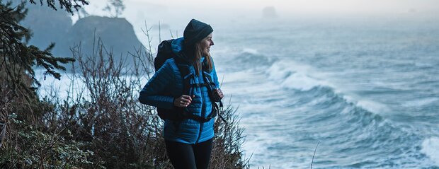 Kvinne i Stormtech turjakke med ryggsekk nyter utsikten ved kysten