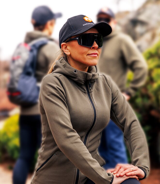 Kvinne iført svart caps med oransje logo, solbriller og en olivengrønn hettejakke. Hun strekker ut etter trening, omgitt av en gruppe mennesker kledd i lignende klær, noen med ryggsekker. Caps med logo er et populært valg for bedriftsprofilering og utendørs aktiviteter. Bakgrunnen er lett uskarp og viser stein og grønn vegetasjon.