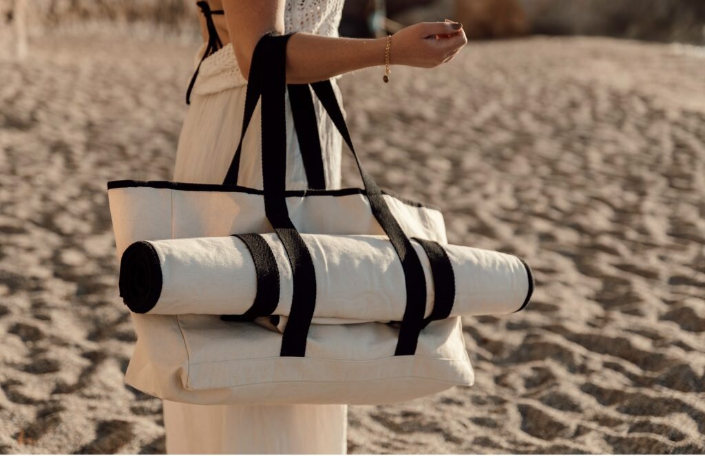 Kvinne i hvitt antrekk på en strand med en Vinga-strandbag og en sammenrullet matte festet til den.