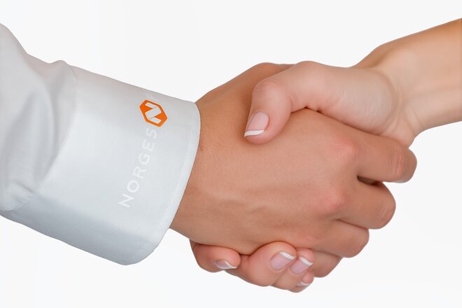 Profesjonelt håndtrykk mellom to personer, hvor den ene har på seg en hvit skjorte med trykket firmalogo. Perfekt bedriftsantrekk for tillitsskapende møter.