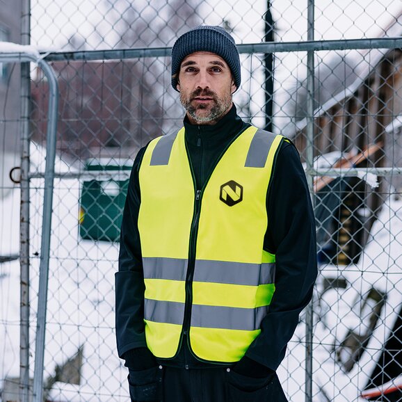 Mann iført en gul refleksvest med grå refleksbånd og logo i front, kombinert med en mørk jakke og lue. Han står utendørs på et industriområde med snø i bakgrunnen. Perfekt for arbeidsklær og synlighet på byggeplasser.
