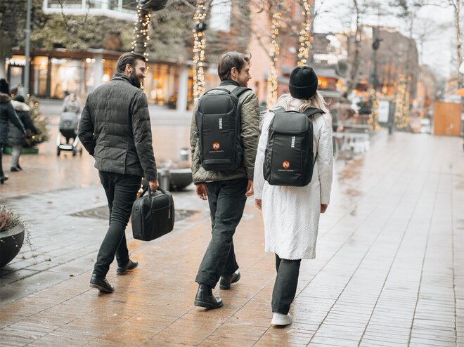Personer iført stilrene svarte ryggsekker med firmalogo, på en urban handlegate med julebelysning. Perfekt profileringsprodukt for bedrifter, events og daglig bruk.