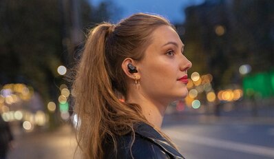 Kvinne med langt hår og Urban Vitamin trådløse ørepropper i øret, poserer i en urban kveldsetting med uskarpe bylys i bakgrunnen.