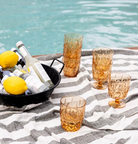 Sagaform amberfargede glass med mønstret overflate plassert på en stripete piknikduk ved bassenget, sammen med en emaljert Sagaform serveringsbolle fylt med sitroner og drikkevarer. Ideell for sommer og utebruk.