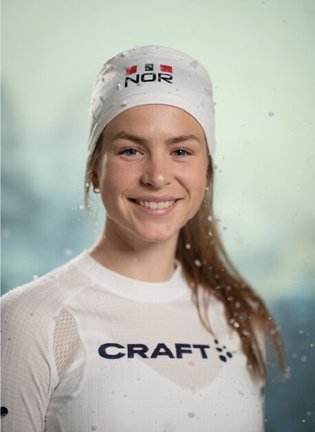 Smilende kvinne i hvit Craft-lue og -trøye, med norsk flagg, fotografert i snøvær.