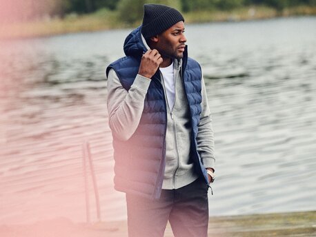 Mann ved en innsjø iført en mørk boblevest, grå hettegenser og svart lue.