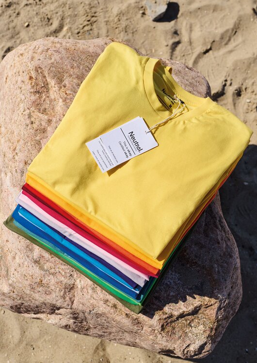 Neutral økologiske T-skjorter i friske farger stablet på strand – bærekraftige profilklær i sertifisert bomull for bedrift