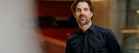 Mann kledd i svart dresskjorte fra YOU Brands, smilende innendørs i et moderne bygg.