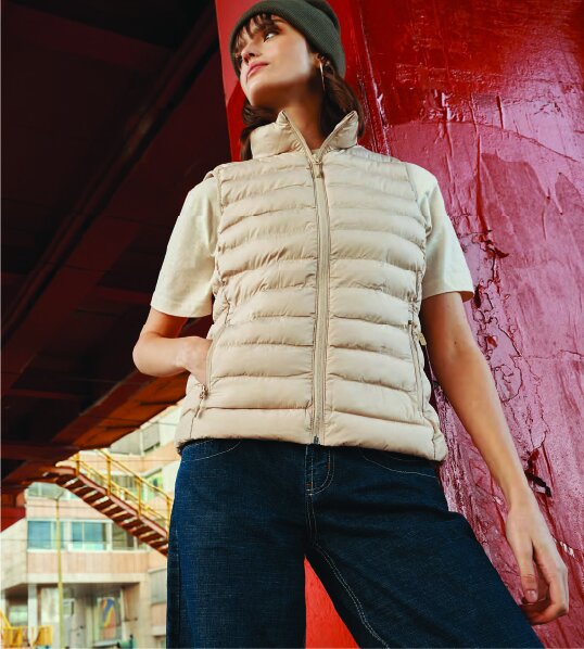 Kvinne iført beige Iqoniq-boblevest og lue, poserer ved en rød søyle i et urbant miljø.