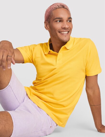 Ung mann med rosa hår iført en gul Roly-pique og lyse shorts, sittende og smilende.