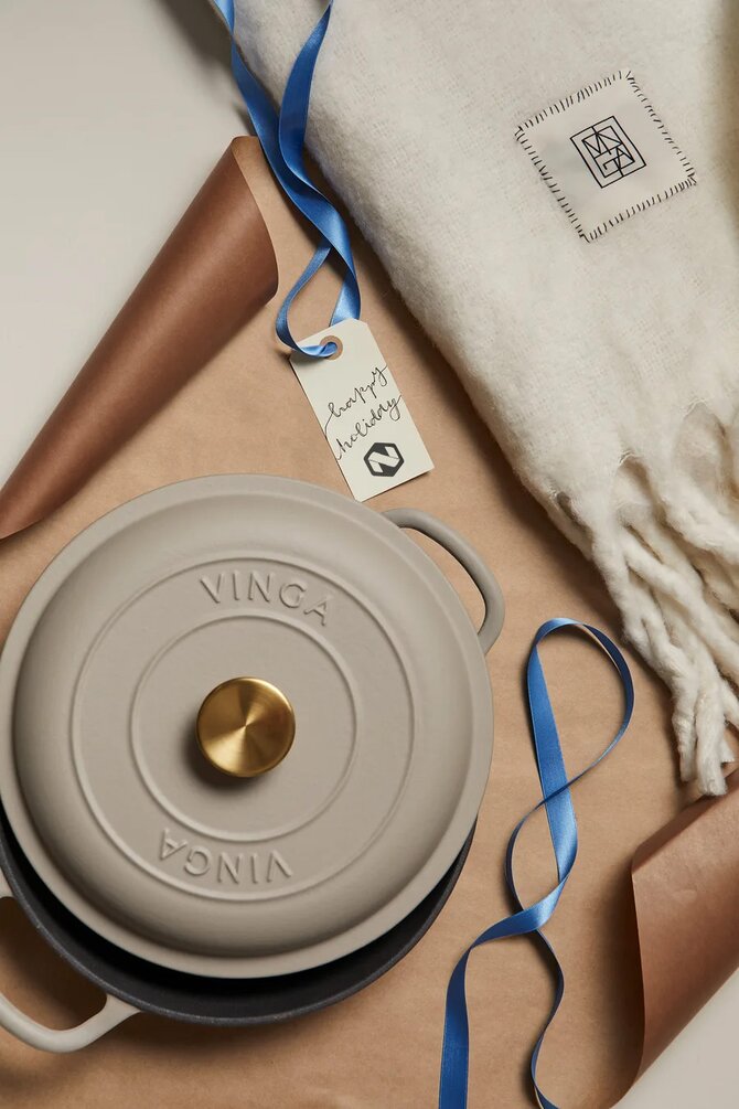 Eksklusiv beige støpejernsgryte med gullfarget håndtak, pakket inn sammen med en myk ullpledd og en håndskrevet gaveetikett – den perfekte firmagaven med kvalitet og omtanke.