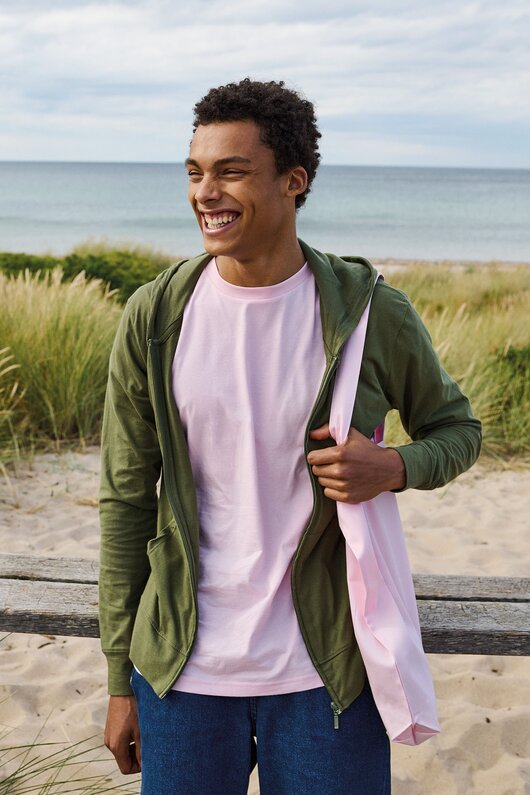 Smilende modell på stranden iført Neutral økologisk T-skjorte og hettejakke – bærekraftige profilklær i sertifisert bomull med naturlig komfort og moderne passform