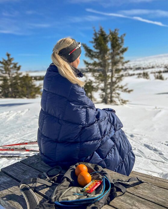 Kvinne i dunpledd nyter påskesolen med appelsin og kvikklunsj – ideell gave for tur- og fritidsaktiviteter.