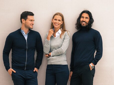 re personer i James Harvest-gensere i blått og grått, smilende foran en lys vegg.