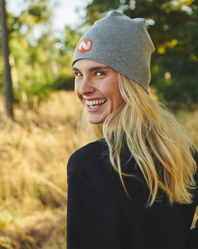 Smilende kvinne med langt blondt hår iført en grå lue med logo, omgitt av solfylt natur med høstlige toner i bakgrunnen. Perfekt profilplagg for kjølige dager utendørs.