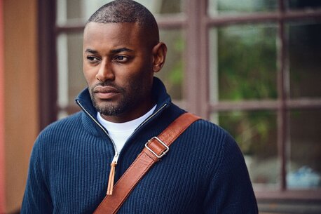 Mann i en mørk, glidelåsgenser fra James Harvest med brun skulderrem, ser til siden.