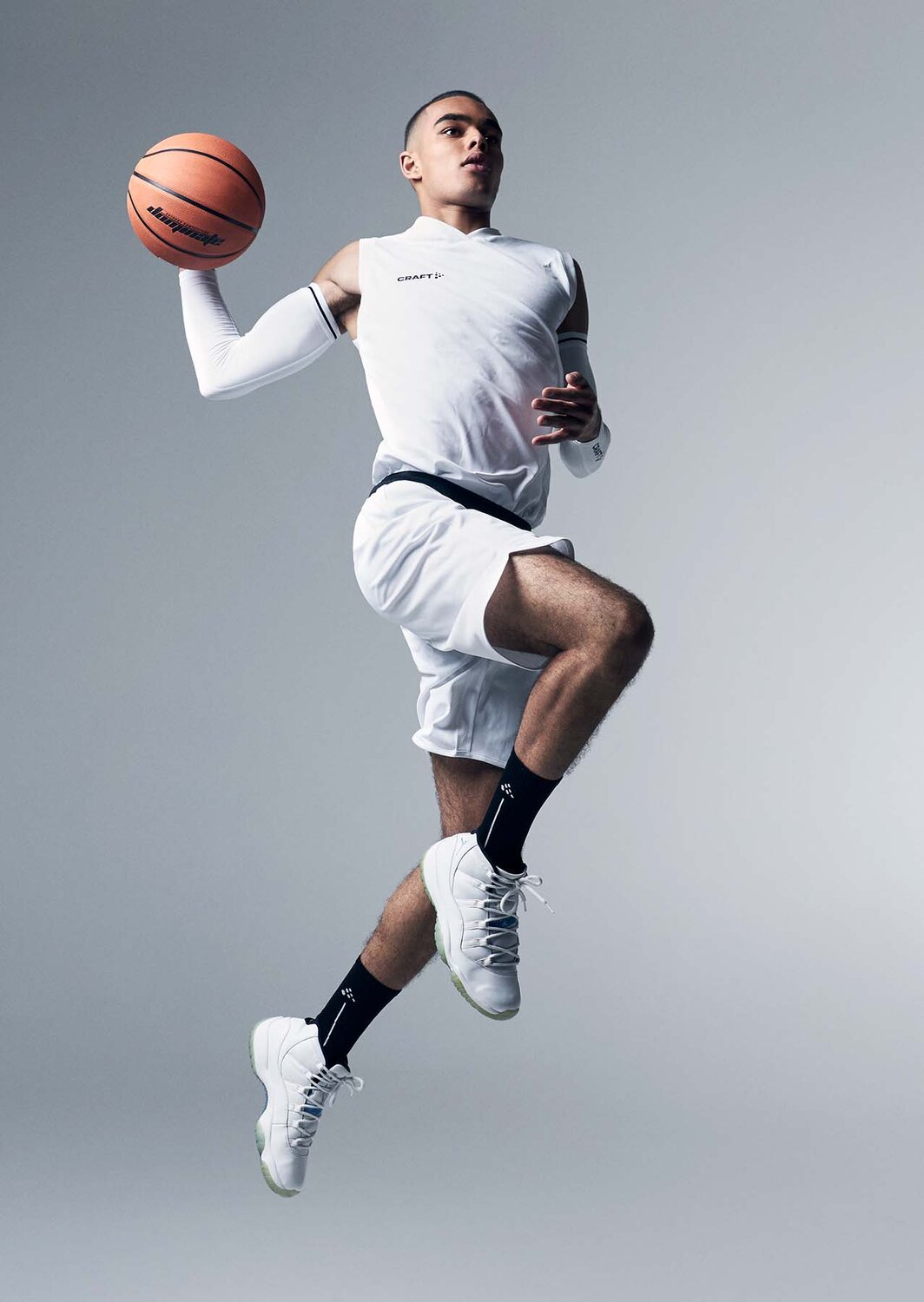 Mann i hvitt Craft Teamwear-basketballantrekk, svevende i et hopp med ballen i hånden.