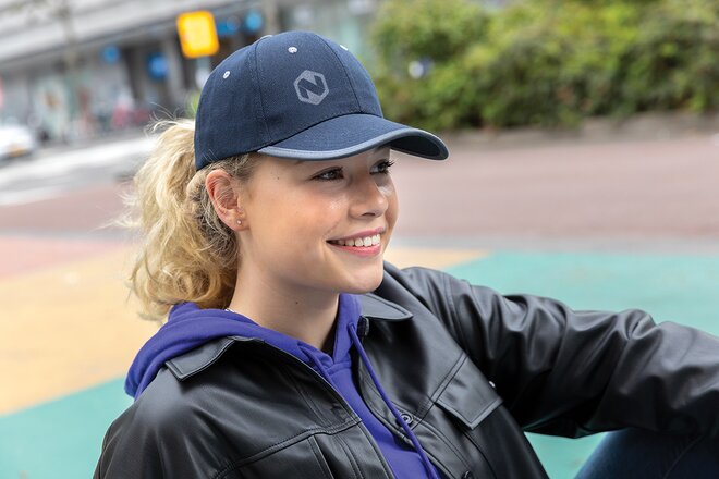 Smilende ung kvinne med krøllete blond hestehale iført en marineblå caps med logo i front. Hun har på seg en svart skinnjakke over en mørkeblå hettegenser. Bildet er tatt utendørs i en urban setting med fargerik asfalt i bakgrunnen.