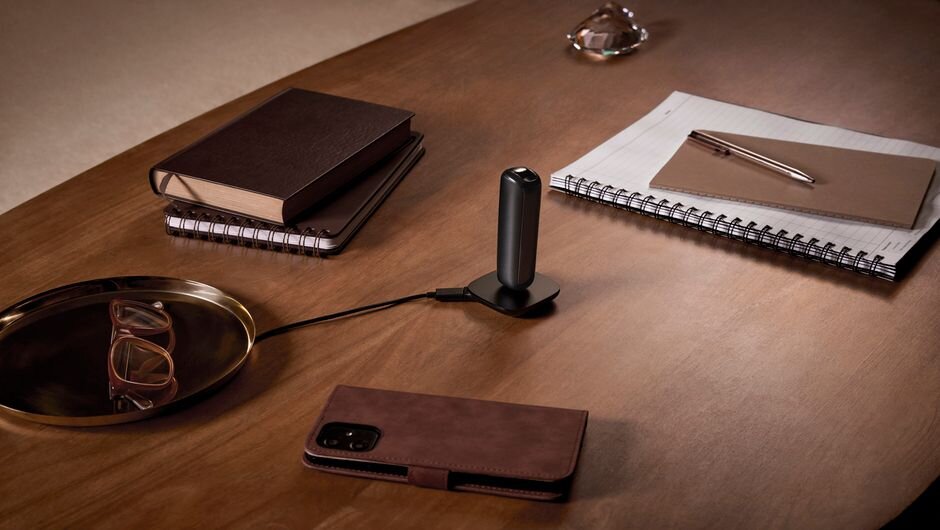 Ploom AURA Jet Black on a charger, surrounded by glasses, notebooks, and a phone with a brown case.