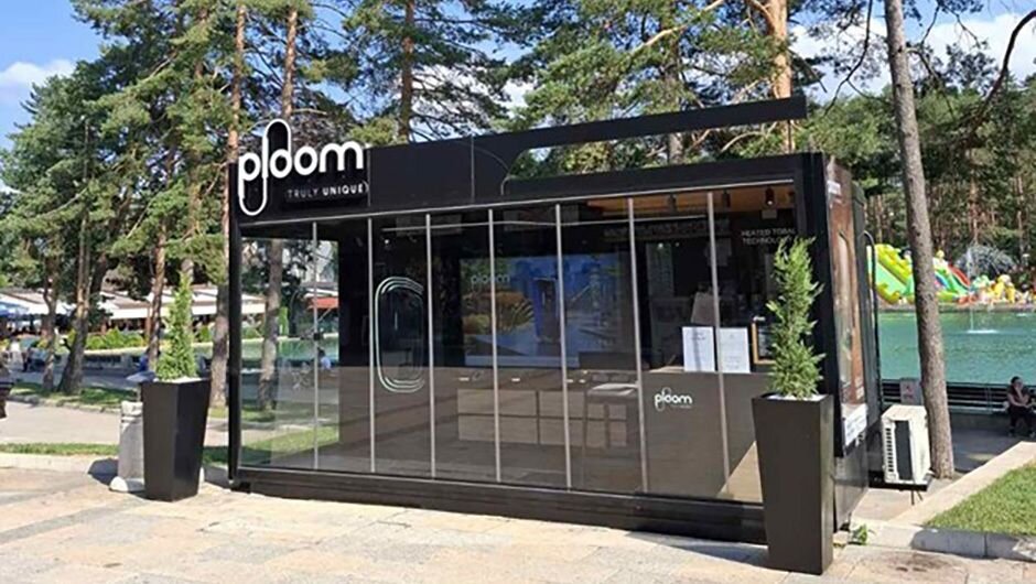 Sleek black Ploom kiosk near pond, amid tall trees, with glass front and promos.