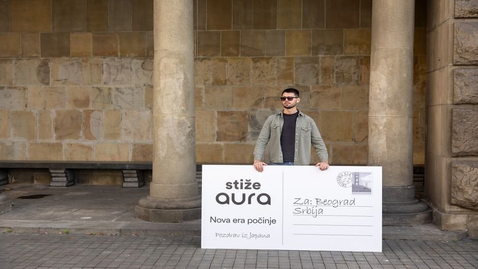 Man with sunglasses holds a Serbian postcard in a modern urban setting with stone columns.
