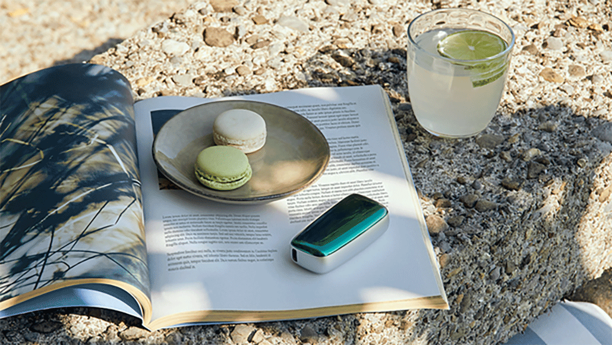 Ploom device on an open magazine, next to macarons and a glass of lemonade on a stone table.