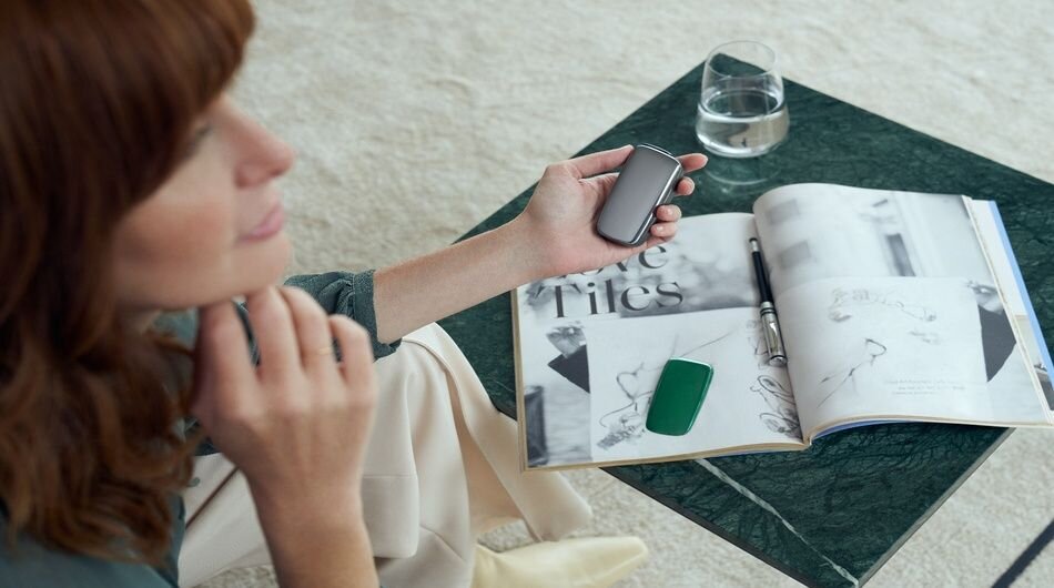 Woman sitting by green marble table holding Ploom X Advance, with magazine, pen and glass of water on table.