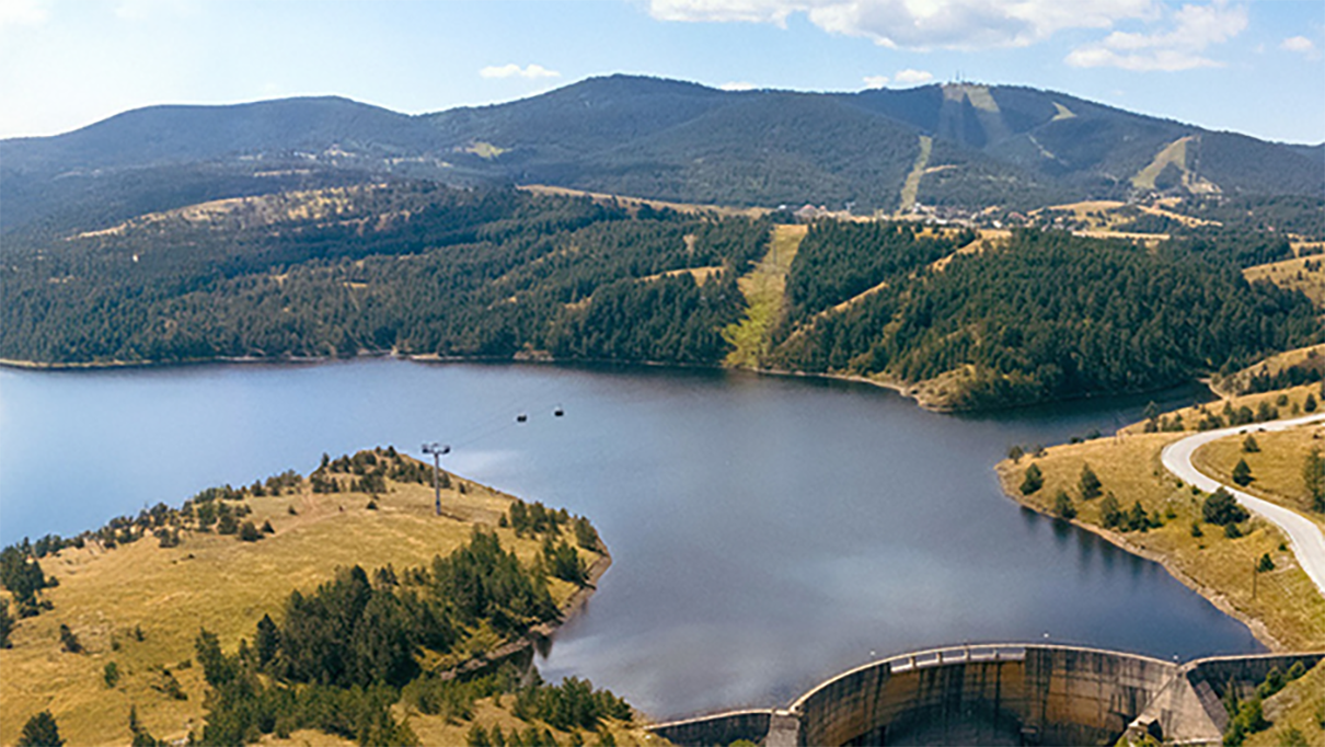 Calm lake with forested hills, blue sky, road and dam in the foreground.