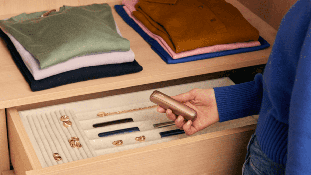 A person holds a Ploom AURA Rose Gold device with a tidy wardrobe and covers in view.