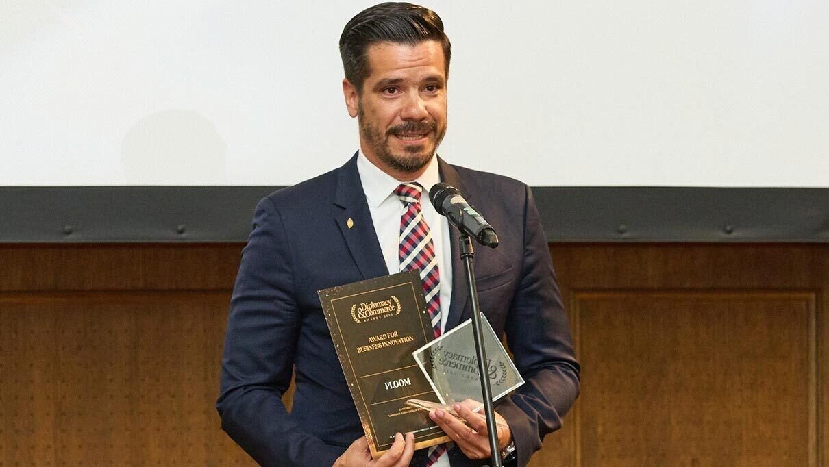 Osoba u odelu drži dve nagrade i stoji za mikrofonom ispred ekrana.