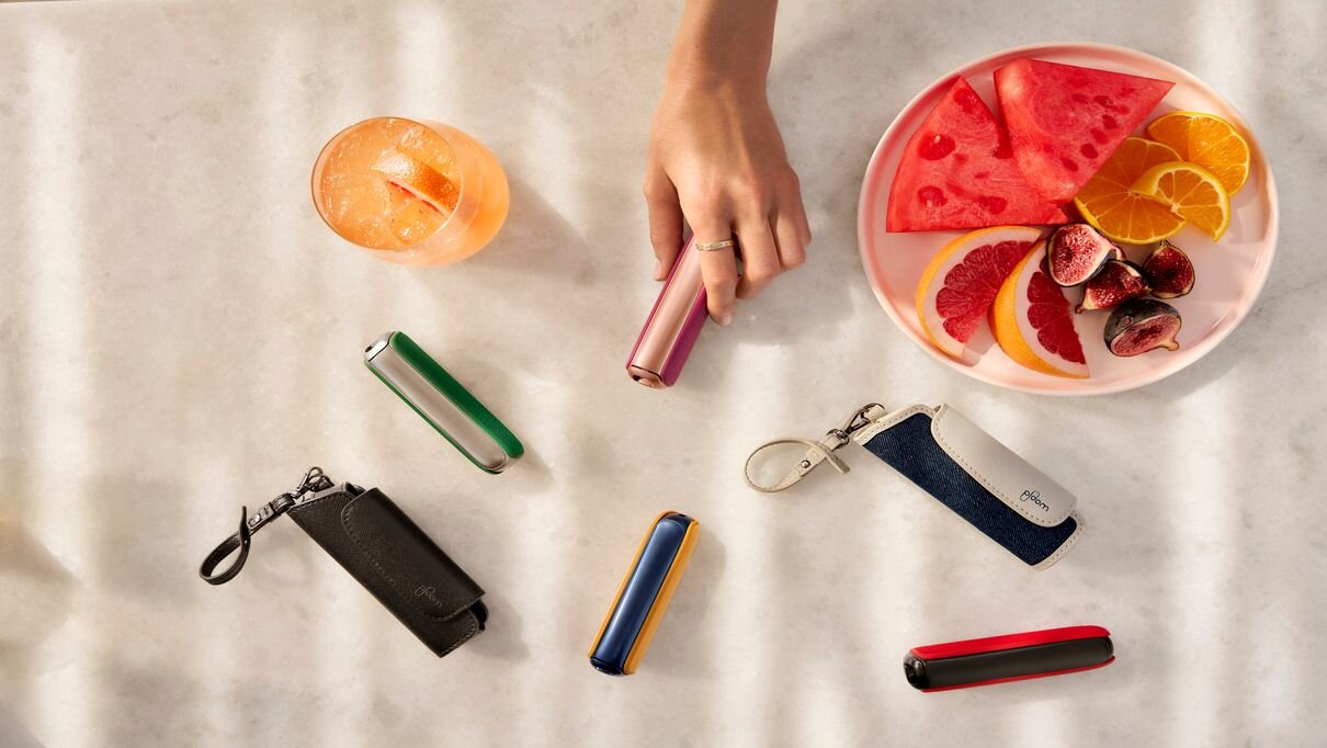 Ploom AURA uređaji u svim bojama okruženi futrolama, tanjirom sa voćem i osvežavajućim pićem.